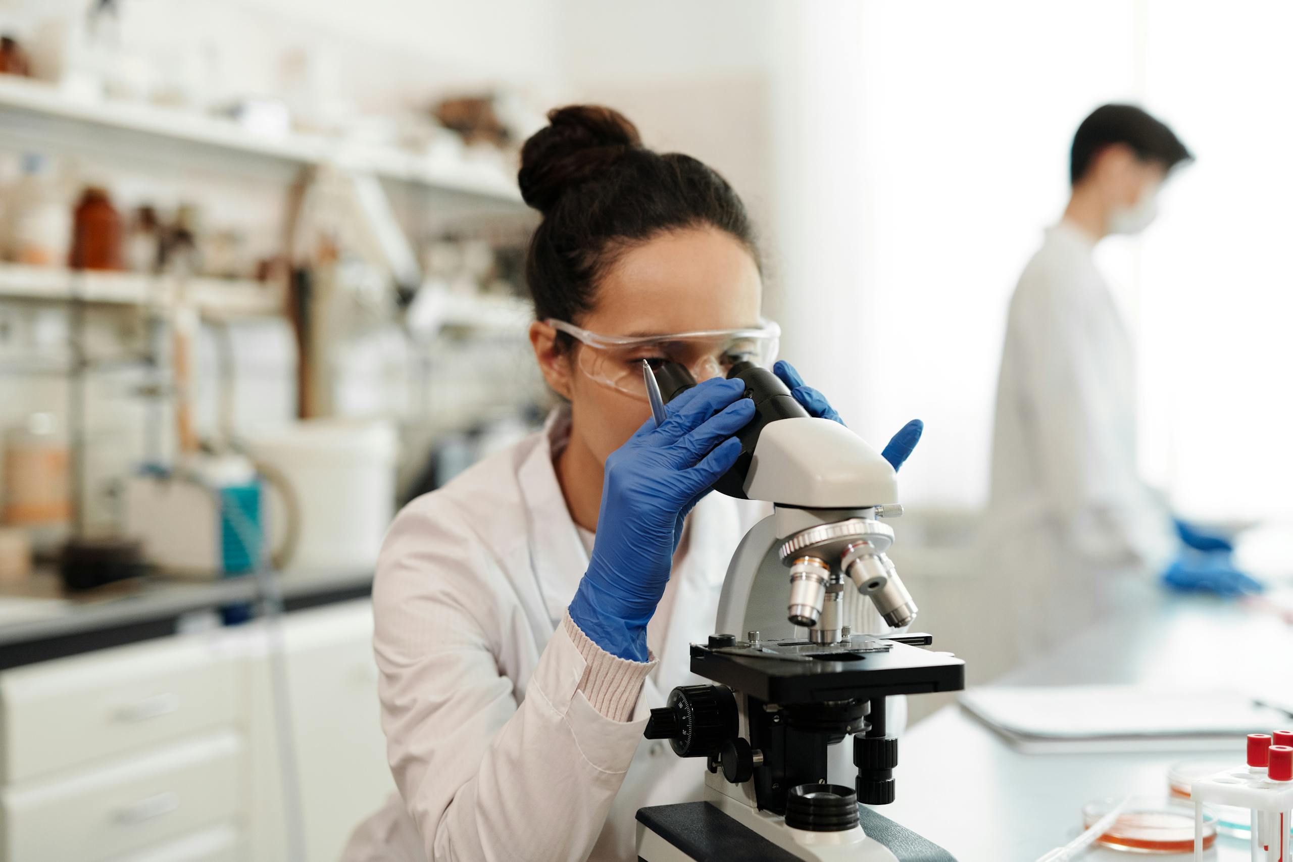 A female scientist in protective gear examines samples through a microscope in a laboratory setting.
